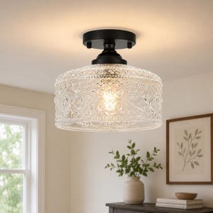 May include: A semi-flush mount ceiling light featuring a matte black metal canopy and a cylindrical glass shade. The clear glass shade is embossed with an intricate, raised scrollwork pattern and a beaded edge, emitting a warm glow.