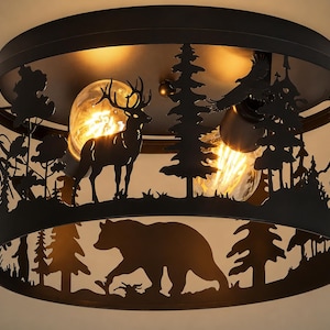 May include: A round flush mount ceiling light fixture with a matte black metal shade featuring a laser-cut forest silhouette. The design includes a standing deer, a walking bear, pine trees, and a flying eagle, illuminated by two warm light bulbs.