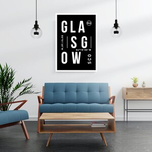 May include: A black and white poster with the word "GLASGOW" in large white letters. The poster also includes the coordinates "N-55.87.15.55" and "W 04.15.00.00". The poster is hanging on a white wall in a living room setting with a blue sofa, a coffee table, and a plant.