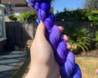 May include: A skein of purple yarn, held up against a sunny background. The yarn's colour shifts from deep indigo to lighter shades of violet, highlighting its texture and suitability for knitting or crochet projects.