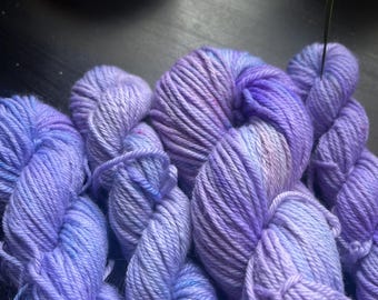 May include: A collection of hand-dyed yarn skeins in various shades of purple and blue. The yarn is arranged together, displaying the texture and colour variations. The lighting accentuates the soft, fluffy texture of the yarn, ideal for knitting or crochet projects.