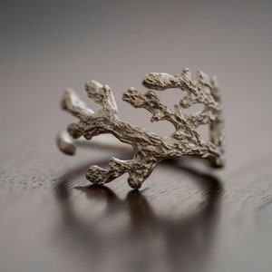 Puede incluir: Un anillo de plata con un diseño delicado e intrincado que se asemeja a una rama o coral. El anillo está hecho de un metal liso y pulido y tiene un aspecto natural y orgánico.