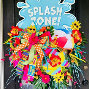 May include: A colorful summer wreath with a blue "SPLASH ZONE!" sign. The wreath features a beach ball, sunglasses, and smiling suns. Bright ribbons, flowers, and greenery complete the festive design.