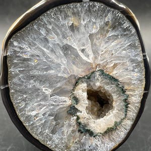 May include: A circular agate geode slice featuring a translucent, crystalline center with jagged white and clear quartz formations. The interior is surrounded by a dark green mineral ring, enclosed by a smooth, polished black and white banded outer edge.