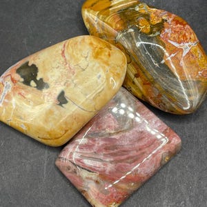 Puede incluir: Tres piedras pulidas en tonos amarillos, naranjas, rojos y grises. Las piedras tienen un acabado liso y brillante y formas variadas, incluyendo ovaladas y de diamante. Las piedras probablemente se utilizan con fines decorativos o como objetos de colección.