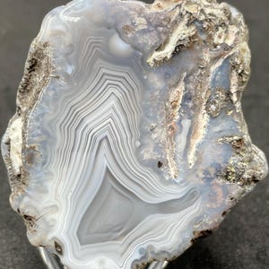 May include: A polished agate slice featuring concentric bands of translucent light blue, white, and gray. The stone has a jagged, rough, and textured outer edge with earthy brown and tan mineral deposits surrounding the smooth, banded center.