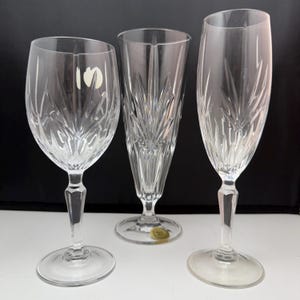 May include: Three clear crystal wine glasses with detailed etched patterns. The set includes a wine glass, a champagne flute, and a larger stemmed glass. Each glass features a distinct shape and design, suitable for a formal occasion.