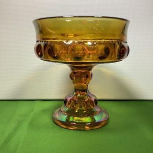 May include: A vintage amber glass pedestal bowl with a textured, iridescent finish. The bowl has a wide, round base and a raised, circular center.