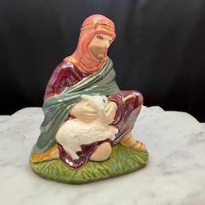 May include: A ceramic figurine of a shepherd kneeling and holding a white lamb. The shepherd is wearing a red robe with a green cloak and a pink turban with gold trim.