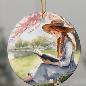 May include: A round ceramic ornament featuring a watercolor illustration of a girl with red hair sitting under a blooming tree and reading a book.