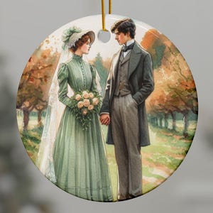 May include: A round ceramic ornament featuring a watercolor illustration of a couple in wedding attire standing in a forest setting. The bride is wearing a white dress with a green sash and a veil. The groom is wearing a black suit and a white shirt. The background is a forest with trees and foliage.
