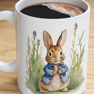 Può includere: Tazza da caffè in ceramica bianca con un coniglio marrone che indossa una giacca blu e sta in piedi in un campo di erba verde e fiori viola.