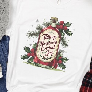 Può includere: T-shirt bianco con grafica di una bottiglia di cordiale al lampone e la scritta "Tidings of Raspberry Cordial and Joy". Il design include agrifoglio e bacche, evocando un tema festivo.
