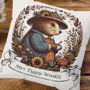 Op de afbeelding: Een wit kussen met een kleurrijke illustratie van een egel die een hoed draagt en een kopje vasthoudt. De illustratie is omringd door bloemen en bladeren. De tekst "Mrs Tiggy-Winkle" is afgedrukt op een banner onder de egel.