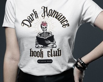 May include: White t-shirt with a graphic of a skeleton reading a book, with the text "Dark Romance Book Club." The design includes a heart and rays. The shirt is worn with black leather pants and bracelets.