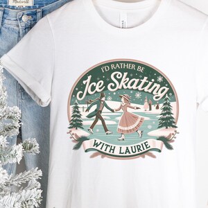 以下が含まれることがあります： 「I'd Rather Be Ice Skating with Laurie」と書かれたグラフィックがプリントされた白いTシャツ。グラフィックは、冬のシーンでスケートをするカップルを描いています。青いジーンズと小さなクリスマスツリーが背景にあります。