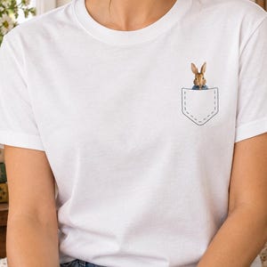 May include: A white short-sleeved t-shirt featuring a graphic of a brown rabbit wearing a blue jacket peeking out from a printed black outline of a chest pocket on the left side. The shirt has a crew neckline and a standard fit.