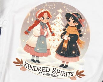 May include: White t-shirt featuring a winter scene with two cartoon girls and the text "Kindred Spirits at Christmas." The design includes a Christmas tree and falling snow. The shirt is displayed with a gray beanie, jeans, and a coffee cup.