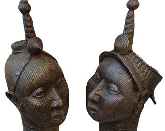 May include: Two dark brown metal sculptures of human heads, each featuring vertical line textures on the face, a tall pointed finial on top, and horizontal ring details around the neck area. Both figures display distinct facial features.
