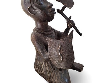Può includere: Scultura in legno marrone scuro di una figura inginocchiata che tiene un martello e uno scudo. Lo scudo ha un motivo a diamante e un disegno a forma di fiamma. La figura ha una testa grande.