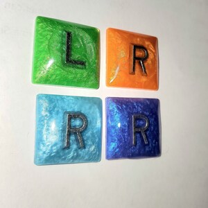 Pode incluir: Quatro azulejos quadrados de resina com glitter embutido na resina. Cada azulejo tem uma letra: L, R, R, R. Os azulejos são verdes, laranja, azuis e roxos.