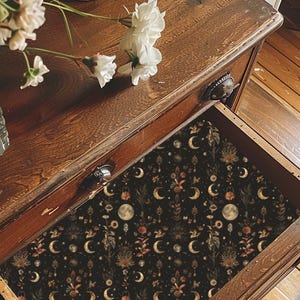 Op de afbeelding: Een houten ladekast met een open lade bekleed met donker papier met een herhalend patroon van manen, sterren en botanische elementen. Een doorzichtige glazen vaas gevuld met witte bloemen staat op het bovenblad van het houten meubelstuk.
