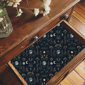 Op de afbeelding: Een donkerhouten ladekast met een open lade bekleed met zwart patroonpapier met botanische illustraties, paddenstoelen en verschillende maanfasen. Een doorzichtige glazen vaas gevuld met witte bloemen staat op het bovenste oppervlak.