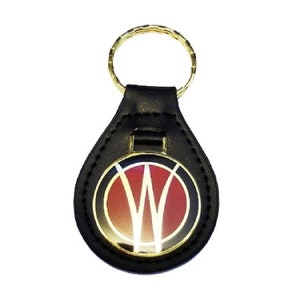 Puede incluir: Llavero de cuero negro con un emblema circular dorado que presenta un logotipo estilizado "W" en rojo y blanco.