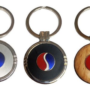 Könnte beinhalten: Drei Schlüsselanhänger mit einem rot-weiß-blauen Logo. Die Schlüsselanhänger sind aus silbernem Metall, schwarzem Metall und Holz.