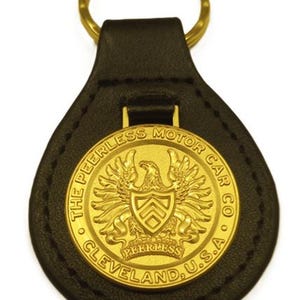 May include: A black leather keychain with a gold-colored metal emblem. The emblem features an eagle and the text "THE PEERLESS MOTOR CAR CO. CLEVELAND, U.S.A.". A gold-colored ring is attached to the top.