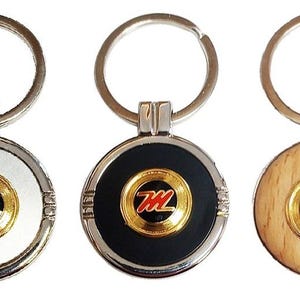 May include: Three keychains with a silver ring and a round metal charm. The charms are silver, black, and wood. Each charm has a gold-colored circle with a red and black logo in the center.