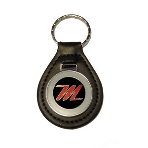 Può includere: Portachiavi in ​​pelle nera con un cerchio di metallo argentato che presenta un logo "M" rosso su sfondo nero.