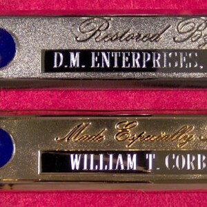 Könnte beinhalten: Zwei rechteckige Plaketten in Silber und Gold mit einem blauen und roten Logo und schwarzem Text. Die obere Plakette lautet "Restauriert von D.M. ENTERPRISES, INC." Die untere Plakette lautet "Speziell angefertigt für WILLIAM T. CORBIN."