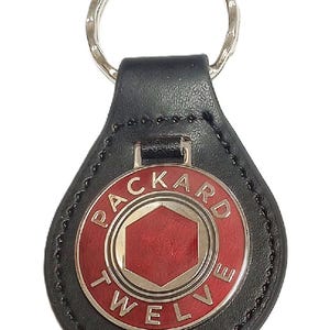 May include: Black leather keychain with a silver key ring. The keychain features a red and silver emblem with the text "PACKARD TWELVE". The emblem has a red hexagon in the center.