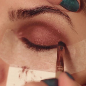 Könnte beinhalten: Nahaufnahme eines Auges, auf das mit einem Make-up-Pinsel Lidschatten aufgetragen wird. Das Auge ist geschlossen und die Person trägt ein weißes Augenpflaster.