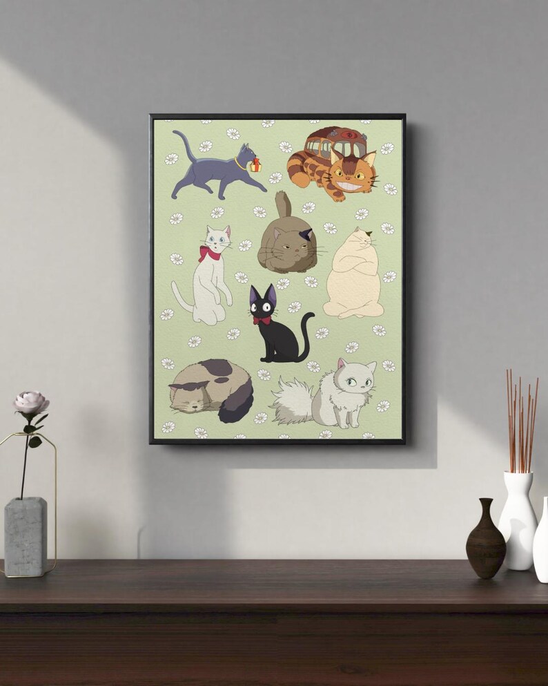 May include: A framed print featuring cartoon cats on a light green background with white daisy accents. The cats are in various poses and colors, including gray, white, black, and orange. The print has a black frame and is displayed on a dark wooden surface.