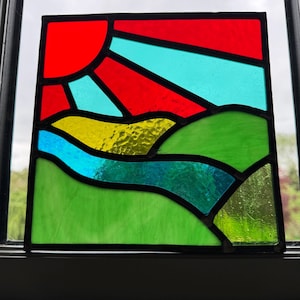 May include: A stained glass window depicting a sunset over rolling hills. The sun is red and orange, with yellow and blue rays. The hills are green, with a blue river winding through them.