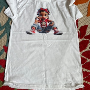 May include: White t-shirt with a cartoon image of a girl wearing a red shirt, blue jeans, and red shoes. The girl is holding a cell phone and has a red bow in her hair. The text "BOOP" is on her shirt.