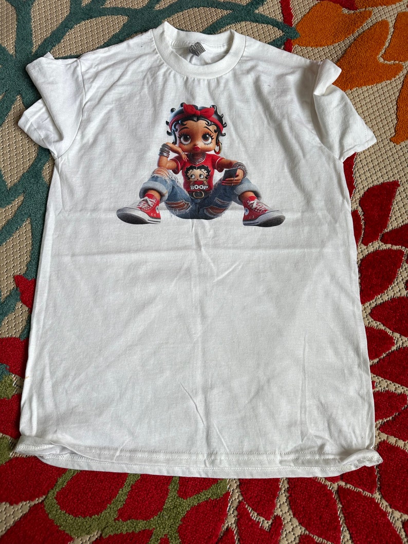 May include: White t-shirt with a cartoon image of a girl wearing a red shirt, blue jeans, and red shoes. The girl is holding a cell phone and has a red bow in her hair. The text "BOOP" is on her shirt.