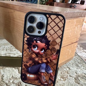 May include: A phone case featuring a cartoon character with large eyes, wearing a plaid shirt and blue jeans. The case has a brown diamond pattern background and a black border. The phone's camera lenses are visible.