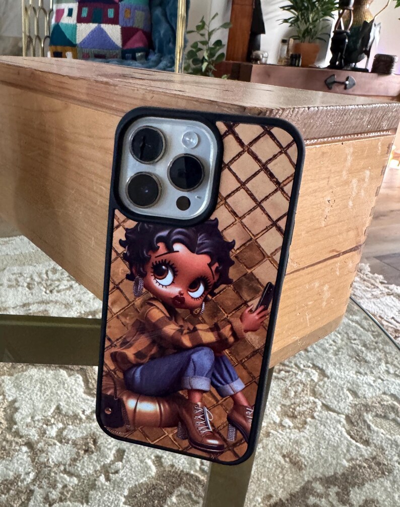 May include: A phone case featuring a cartoon character with large eyes, wearing a plaid shirt and blue jeans. The case has a brown diamond pattern background and a black border. The phone's camera lenses are visible.