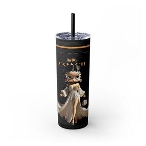 May include: Black tumbler with a black straw featuring a cartoon Betty Boop character in a white dress, holding a purse. The tumbler has the Coach logo and a gold band around the top.