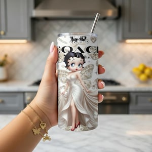 May include: A stainless steel tumbler featuring a design with the Coach logo and a cartoon image of Betty Boop in a white gown. The tumbler has a silver straw and a white and beige background.
