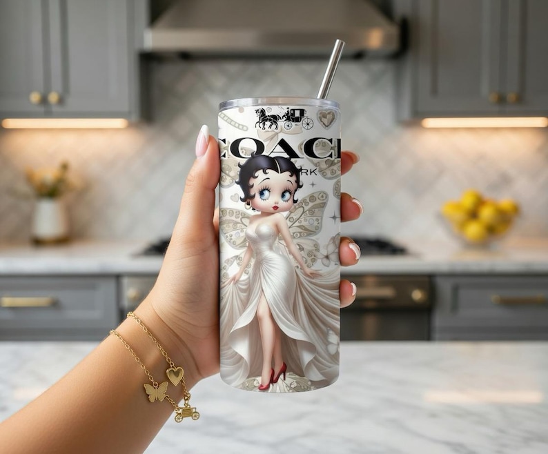May include: A stainless steel tumbler featuring a design with the Coach logo and a cartoon image of Betty Boop in a white gown. The tumbler has a silver straw and a white and beige background.