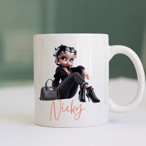 May include: White ceramic mug with a cartoon image of a woman wearing a black jacket, black pants, and black boots. She is holding a black purse and sitting with her legs crossed. The mug has the name "Nicky" written in pink on the bottom.