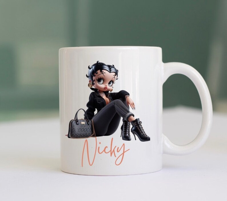 May include: White ceramic mug with a cartoon image of a woman wearing a black jacket, black pants, and black boots. She is holding a black purse and sitting with her legs crossed. The mug has the name "Nicky" written in pink on the bottom.