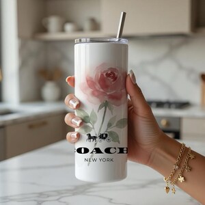 May include: White tumbler with a watercolor rose design and a silver straw. The tumbler features the Coach logo and the words "NEW YORK" in black. The floral design includes pink and green hues.