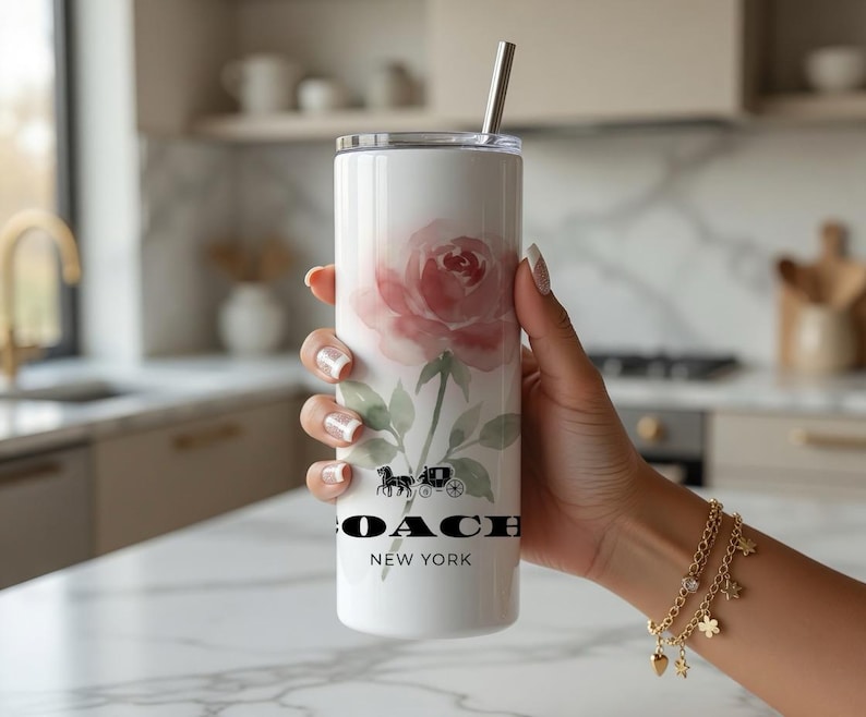 May include: White tumbler with a watercolor rose design and a silver straw. The tumbler features the Coach logo and the words "NEW YORK" in black. The floral design includes pink and green hues.