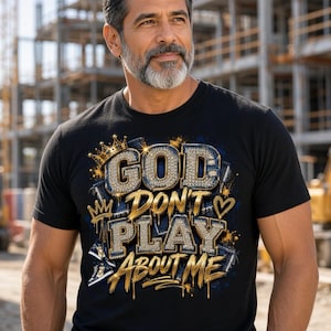 May include: A black t-shirt featuring a graphic design with the text GOD DON'T PLAY ABOUT ME in gold and diamond-textured lettering. The design includes a gold crown, a heart symbol, and blue paint splatters, set against a dark fabric background.