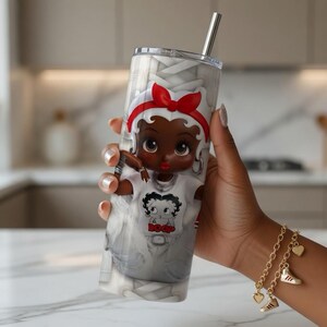 May include: A stainless steel tumbler with a cartoon character design. The character has a red bow and a white shirt. The tumbler has a silver straw and a clear lid. The background is grey and white.
